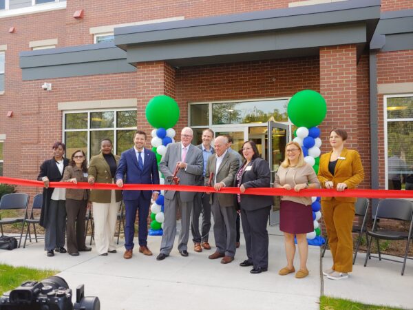 Men and women stand in pose as they hold a large red ribbon in front of a red brick building. Large green balloons mark the sides of the main entrance behind the people.