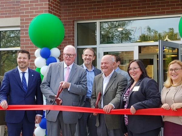 Men and women pose in front of a red brick building holding two large scissors to a large red ribbon. A large green balloon marks the side of the main entrance behind the people.