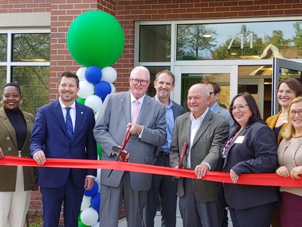 Men and women pose in front of a red brick building holding two large scissors to a large red ribbon. A large green balloon marks the side of the main entrance behind the people.