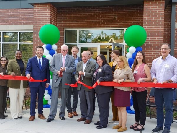 Men and women pose in front of a red brick building holding two large scissors to a large red ribbon. The ribbon has just been cut and a part flutters in the breeze. Two large green balloons mark the side of the main entrance behind the people.