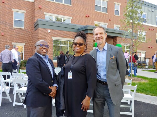 A shorter bald black man wearing glasses and a navy suit poses with a black woman wearing sunglasses, a black dress with an ID on a lanyard, and a tall thin white man wearing a grey suit with a light blue shirt. They are all smiling as they stand in front of a red-brick building.