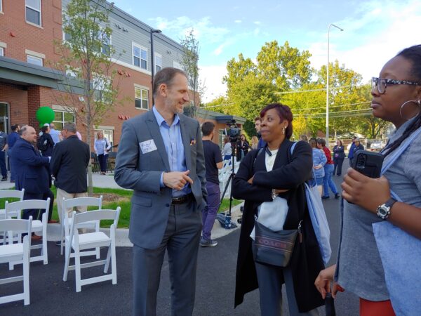 A tall thin white man with brown hair wearing a grey suit and a light blue shirt smiles as he looks at a black woman wearing a dark knee-length jacket over a white shirt. Her arms are crossed as she smiles back.