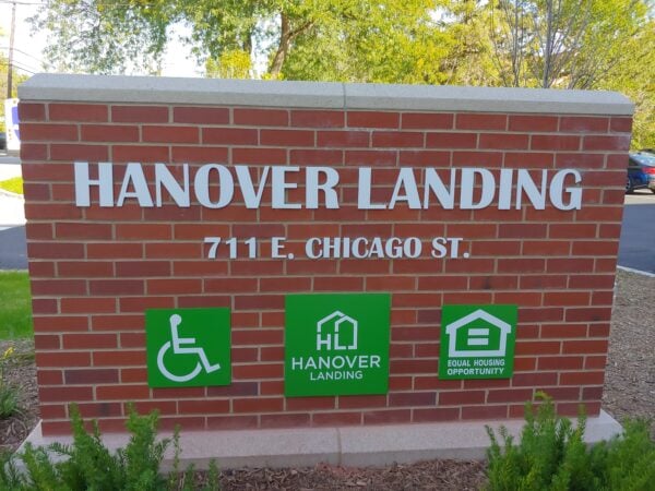 Building signage in red brick. White letters spell out Hanover Landing 711 E Chicago St. Three small green signs squares below the lettering show the Wheelchair icon, the Fair Housing logo, and the Hanover Landing logo.