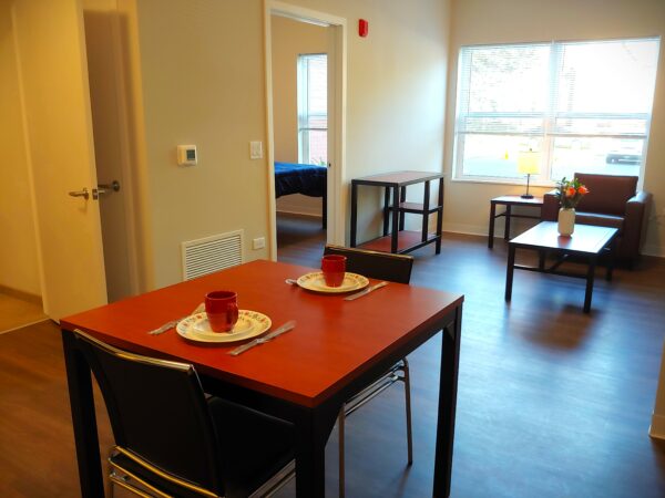 A dine-in kitchen with a small table with two chairs that has two place settings is in the foreground. Wooden floors reflect light from the windows. A glimpse of the bedroom is in the background through an open door.
