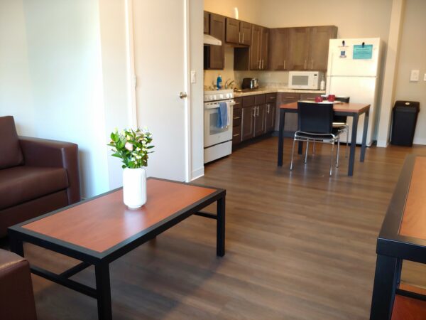 Living room with brown leather couch and chair. A coffee table with a warm wood top and black trim has a vase of fresh flowers. The room's wood floors continue to a dine-in kitchen with a small table with two chairs that has been two place settings.