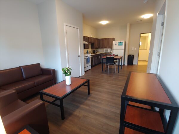 Living room with brown leather couch and chair. A coffee table with a warm wood top and black trim has a vase of fresh flowers. The room's wood floors continue to a dine-in kitchen with a small table with two chairs that has been two place settings.
