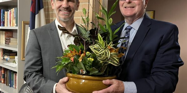 Two men wearing suits stand holding a large pot with plants.