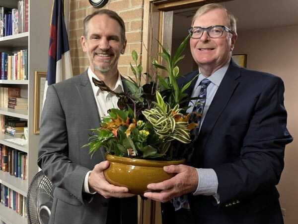 Two men wearing suits stand holding a large pot with plants.