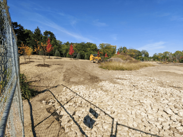 Construction site with rocks in the foreground, a yellow construction vehicle, and several small trees that are red with fall colors.