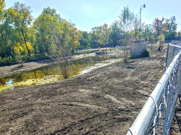 A muddy construction site with a retention pond surrounded by trees and a metal construction fence.