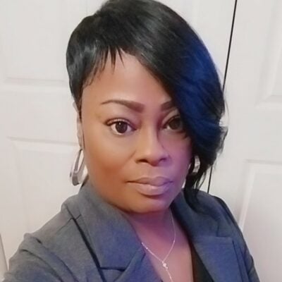 Black woman with short black hair, wearing a grey business jacket, silver hoop earrings and a thin chain necklace.