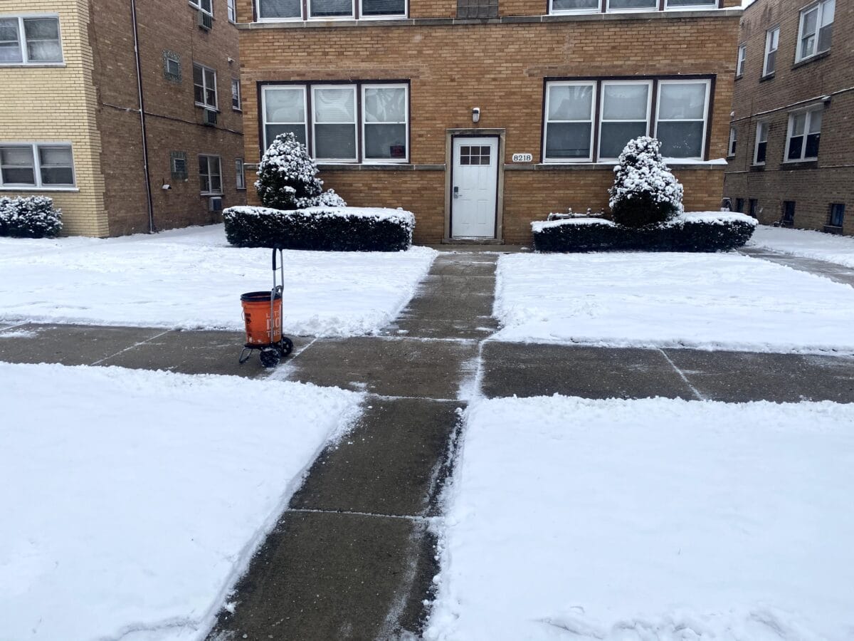 A brick apartment building in winter, 3 inches of snow cover the lawn on both sides of a sidewalk and walkway that has been cleared of snow. A red snowblower sits on the sidewalk.