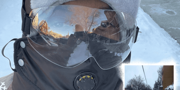 A black man wearing a winter hat, face mask and clear eye guard stands in front of a sidewalk cleared of snow. Snow Warrior is across the top of the picture with small icicles across the top.