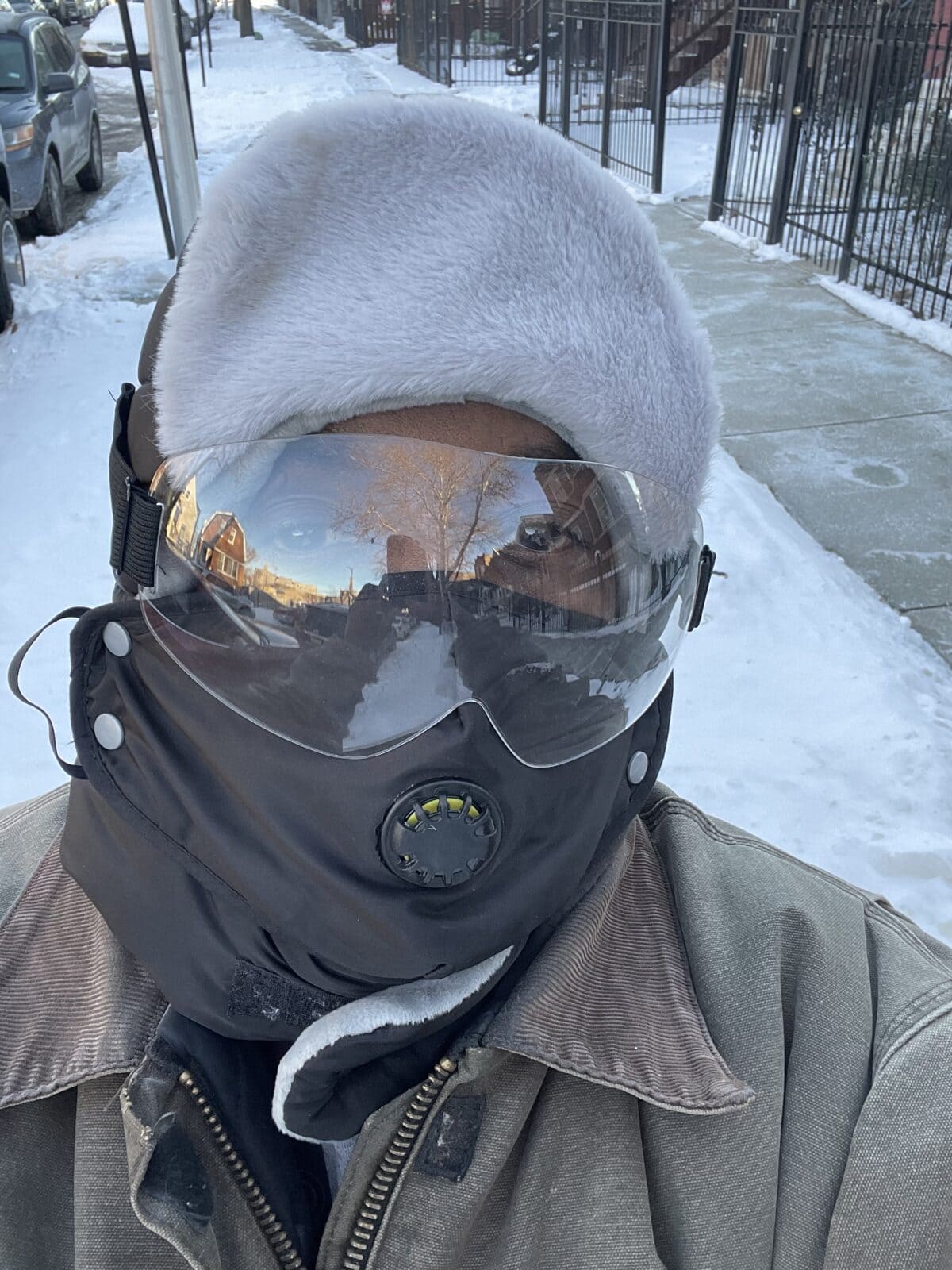 Black man wearing a winter coat and hat, mask, and clear eye guard stands in front of a sidewalk that has been cleared of snow.