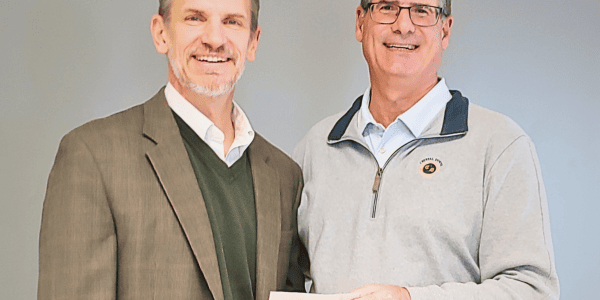 Two men smile as they hold a check. One is wearing a green sweater vest and plaid suit jacket while the other is wearing jersey with a business logo over a white shirt and glasses.