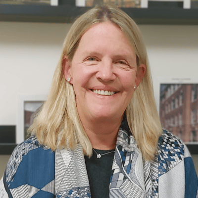 White woman with straight blond hair wearing a blue patterned scarf over a black shirt, earrings and a necklace.