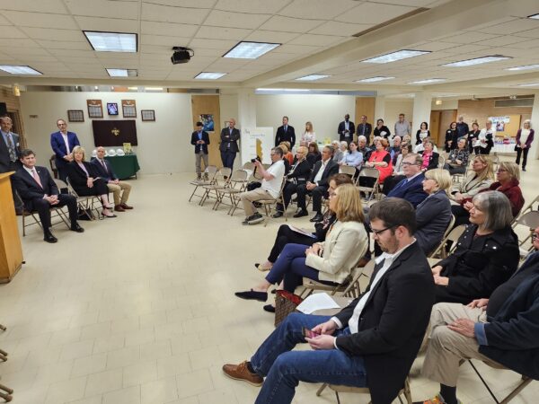 People sitting and standing watching a panel of speakers taking turns speaking at a podium.