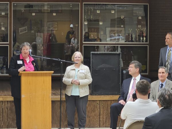 Two women stand a podium while one speaks into a microphone. A white man stands to the side signing and other men and women watch the speaker.