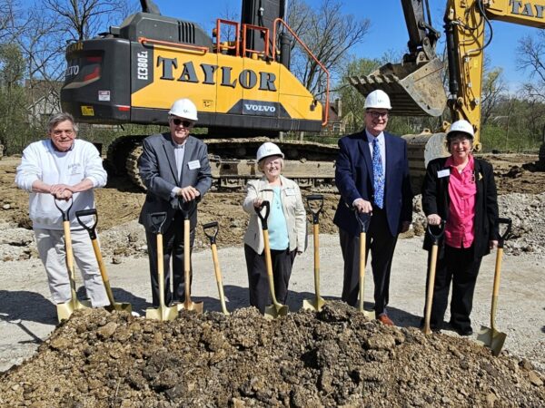 Five men and women wearing white hard hats pose with shovels on a construction site with a large excavator in the background.