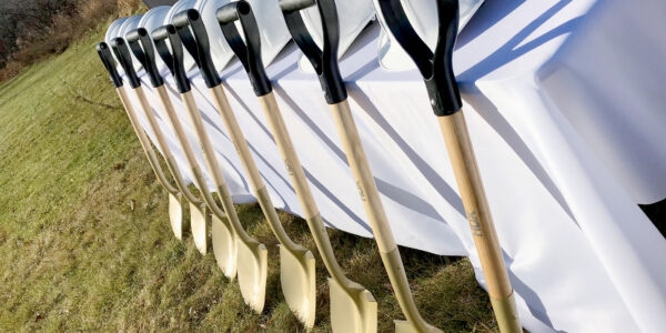 Gold shovels leaning against a table with a white tablecloth and white hard hats aligned on the table.