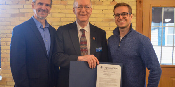Three white men pose. Two are wearing suits and one wearing a blue shirt. The man in the middle is holding a formal document with a gold seal.