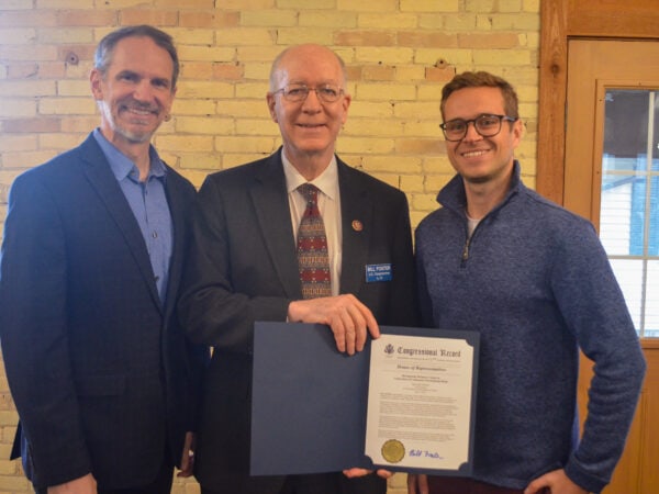 Three white men pose. Two are wearing suits and one wearing a blue shirt. The man in the middle is holding a formal document with a gold seal.