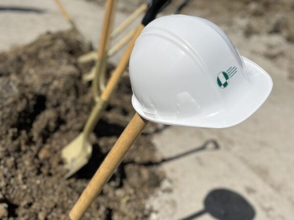 A white hard hat is perched on a golden shovel sticking out of dirt at a construction site with several shovels lined up in the background.