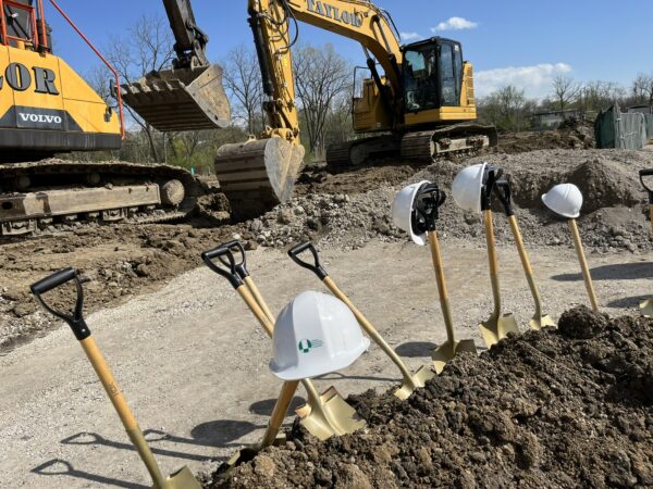 A white hard hat is perched on a golden shovel sticking out of dirt at a construction site with a large excavator in the background.