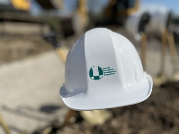 A white hardhat with a green HODC logo with a blurred construction site in the background.