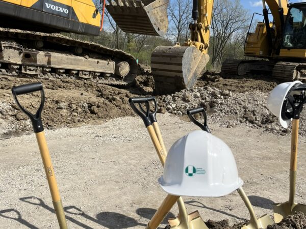 A white hard hat is perched on a golden shovel sticking out of dirt at a construction site with a large excavator in the background.