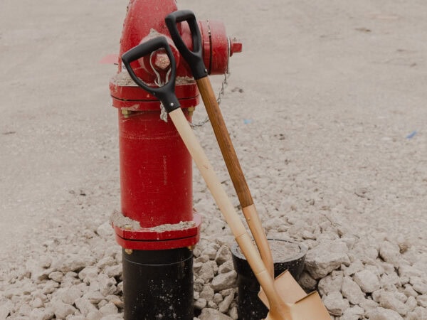 Two cold shovels lean against a red fire hydrant sticking up from a rocky construction site. A construction vehicle and construction materials are visible in the background.