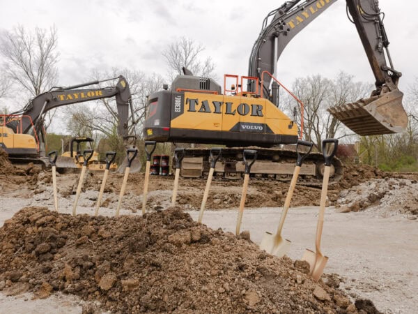 Gold shovels are sticking up from a pile of dirt in front of two large construction excavators.