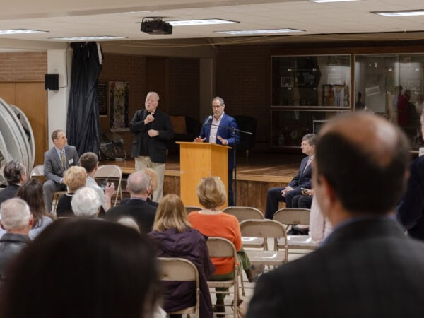 A white man speaks into a mic at a podium as a white man signs next to him. Several men sit next to the podium listening. The back of the heads of the audience are in the foreground.