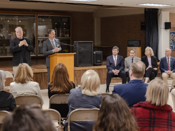 A white man speaks into a mic at a podium as a white man signs next to him. Four men and women sit next to the podium listening. The back of the heads of the audience are in the foreground.