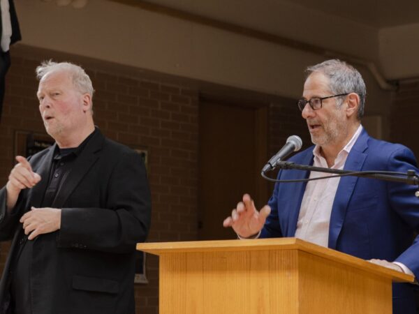 A white man with short salt and pepper hair, wearing a navy blazer and a white shirt speaks into a mic at a podium. Another white man wearing a black blazer over a black shirt and tan pants, is gesturing next him in ASL.