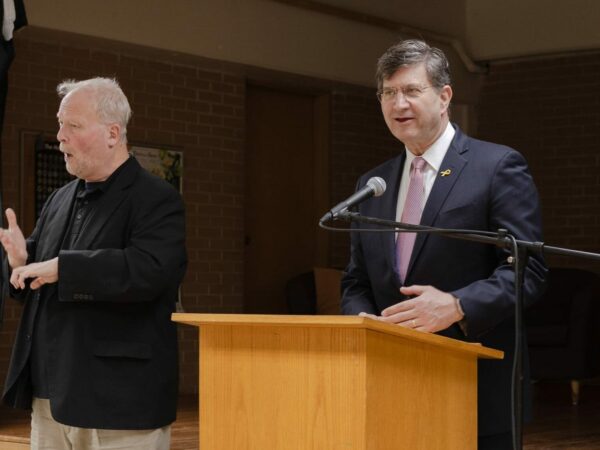 A white man with brown hair, wearing a navy suit with a white shirt and lavender tie speaks into a mic at a podium. Another white man wearing a black blazer over a black shirt and tan pants, is gesturing next him in ASL.