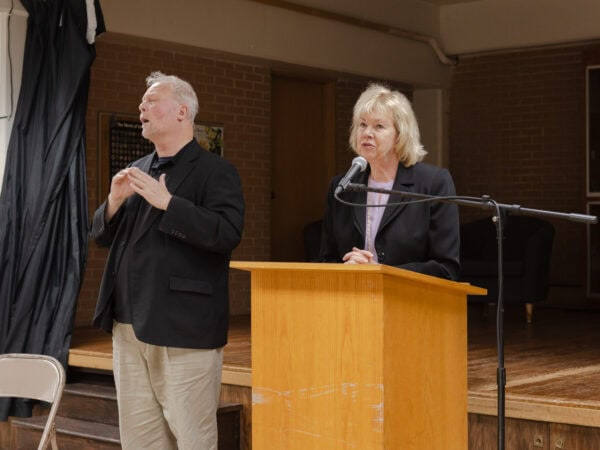 A white woman with shoulder-length blond hair, wearing a black blazer over a light lavender blouse speaks into a mic at a podium. A white man wearing a black blazer over a black shirt and tan pants, is gesturing next him in ASL.