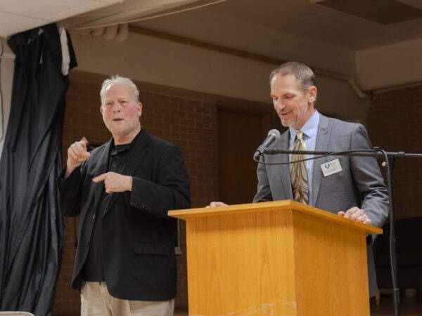 A white man with short salt and pepper hair, wearing a light grey blazer, gold and brown tie, and light blue shirt speaks into a mic at a podium. Another white man wearing a black blazer over a black shirt and tan pants, is gesturing next him in ASL.