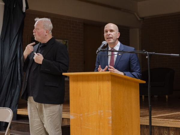 A bald white man wearing a blue blazer, maroon tie, and light blue checkered shirt speaks into a mic at a podium. Another white man wearing a black blazer over a black shirt and tan pants, is gesturing next him in ASL.