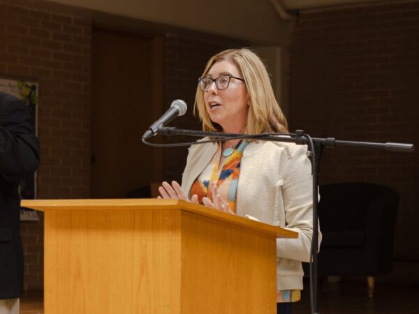 A white woman with shoulder-length blond hair, wearing a light blazer over a muli-colored blouse, speaks into a mic at a podium.