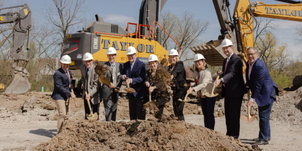 Professionally dressed people wear hard hats and hold shovels of dirt on a construction site with a large construction digger in the background.
