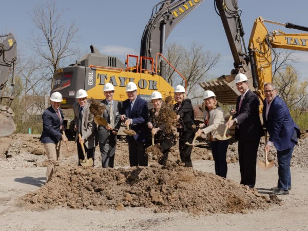 Professionally dressed people wear hard hats and hold shovels of dirt on a construction site with a large construction digger in the background.