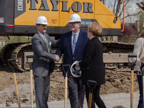 Two men wearing business suits shake hands as they talk with a woman wearing a black outfit. Large construction equipment in the background.