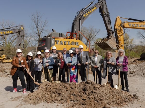 A group of people, some wearing white hard hats pose with shovels on a construction site with large construction vehicles in the background.