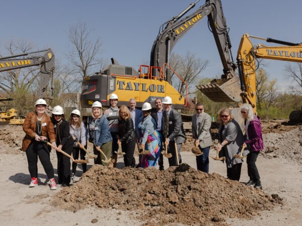 A group of people, some wearing white hard hats are smiling as they shovel dirt on a construction site with large construction vehicles in the background.