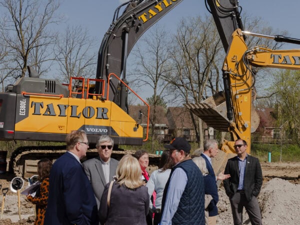 Professionally dressed people stand in front of a large excavator on a construction site.