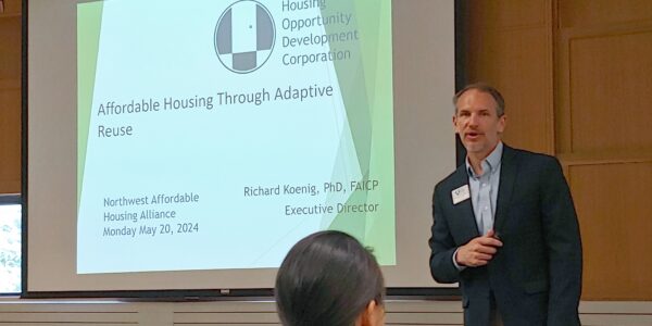 A man wearing a dark blue jacket and light blue button shirt speaks in front of a screen with a projected PowerPoint slide.
