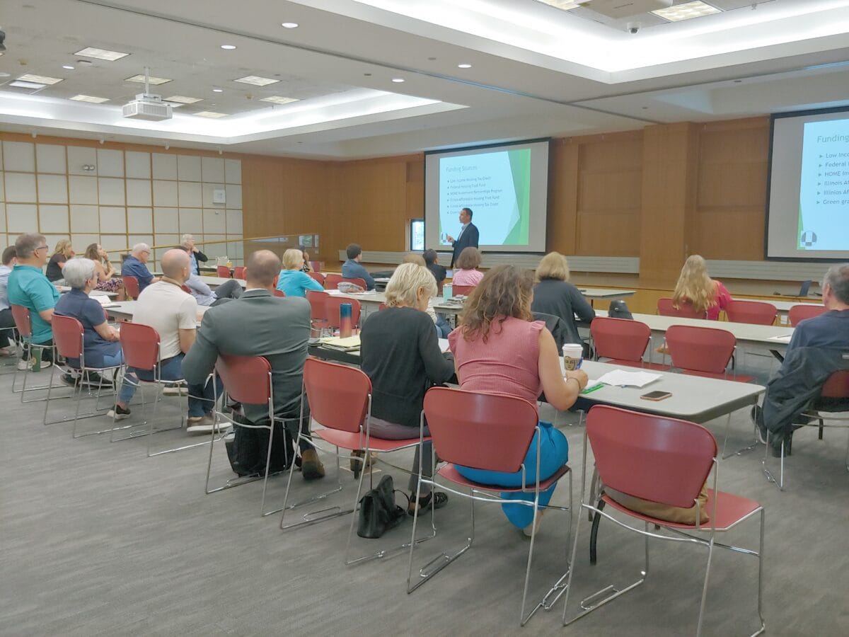 People sitting in a library conference room listening to a man speaking. A Powerpoint is projected on large screens behind him.
