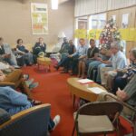 A group of people sitting in chairs in a circle listening to one man talking.