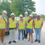 A group of black and white men and women pose together wearing white hard hats and safety vests with a construction site in the background.
